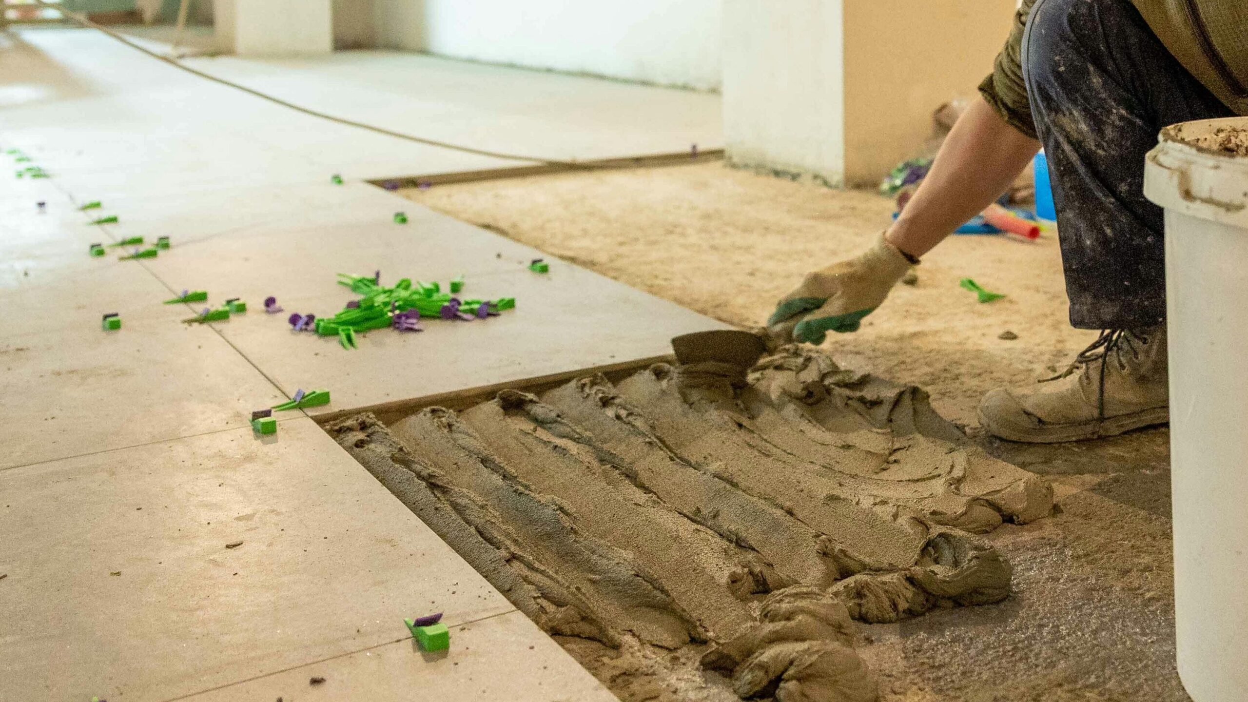 A construction worker lays tiles in an indoor renovation project, wearing safety gear.